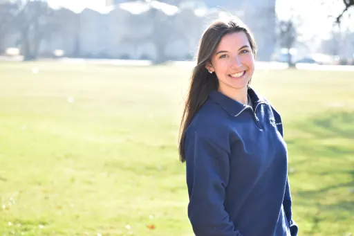 A student in a blue top standing outdoors with a green field and trees in the background