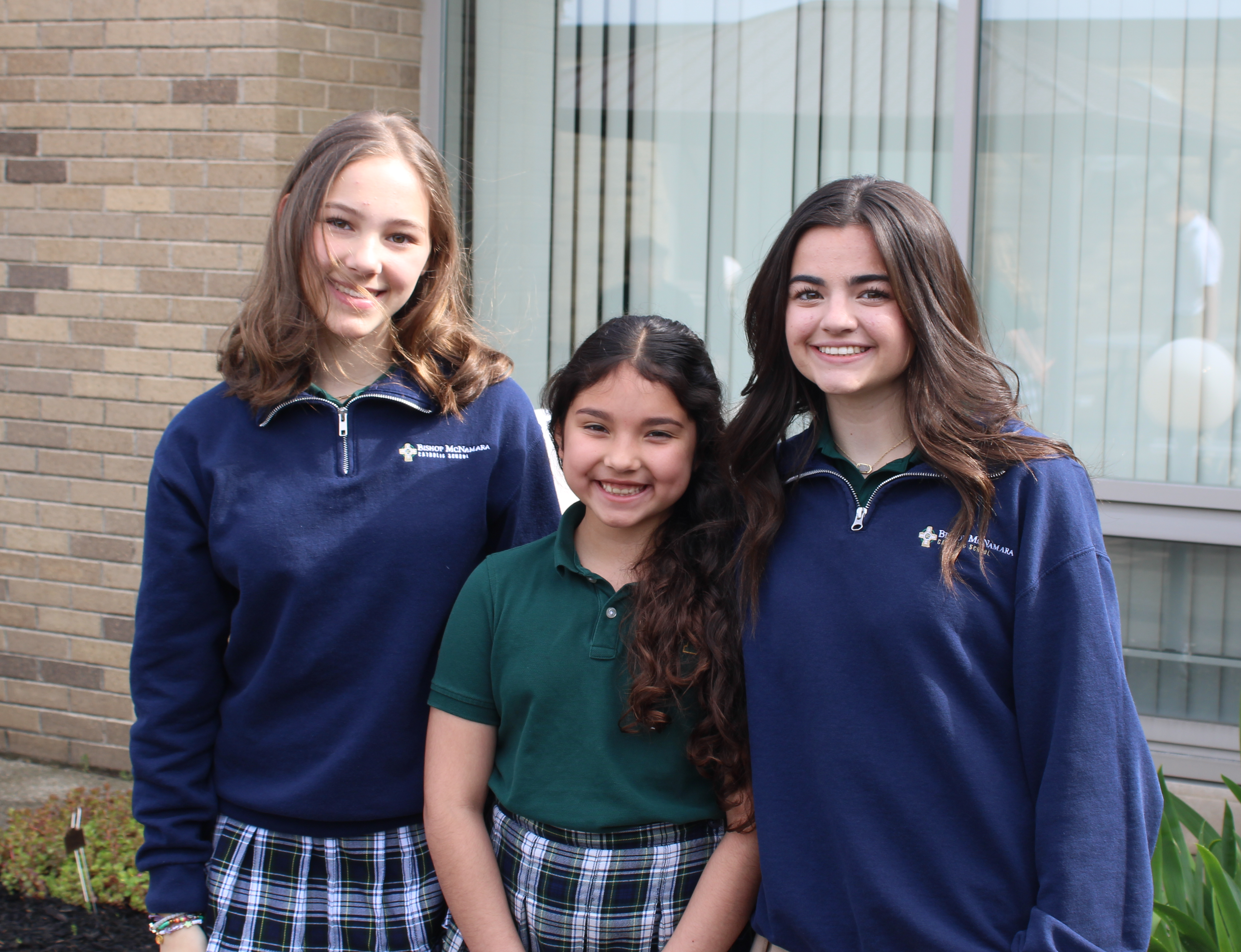 Three students in green polo shirts and khaki shorts standing outside a brick building