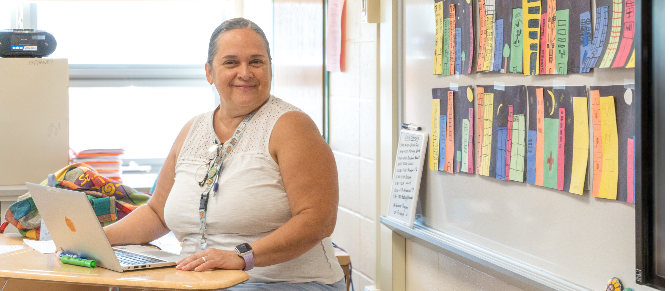 A summer program teacher sits at her desk smiling towards the camera. Student artwork is  prominently dsiplayed  on a board to her left.