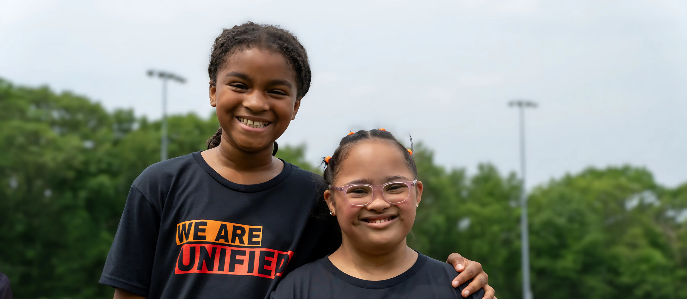 Two students wearing "We are Unified" shirts pose for a group photo outside.