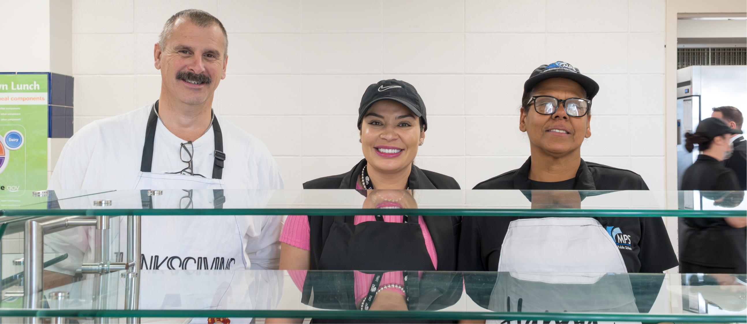 Three people pose behind a glass counter; two women in aprons and a man in a white shirt.