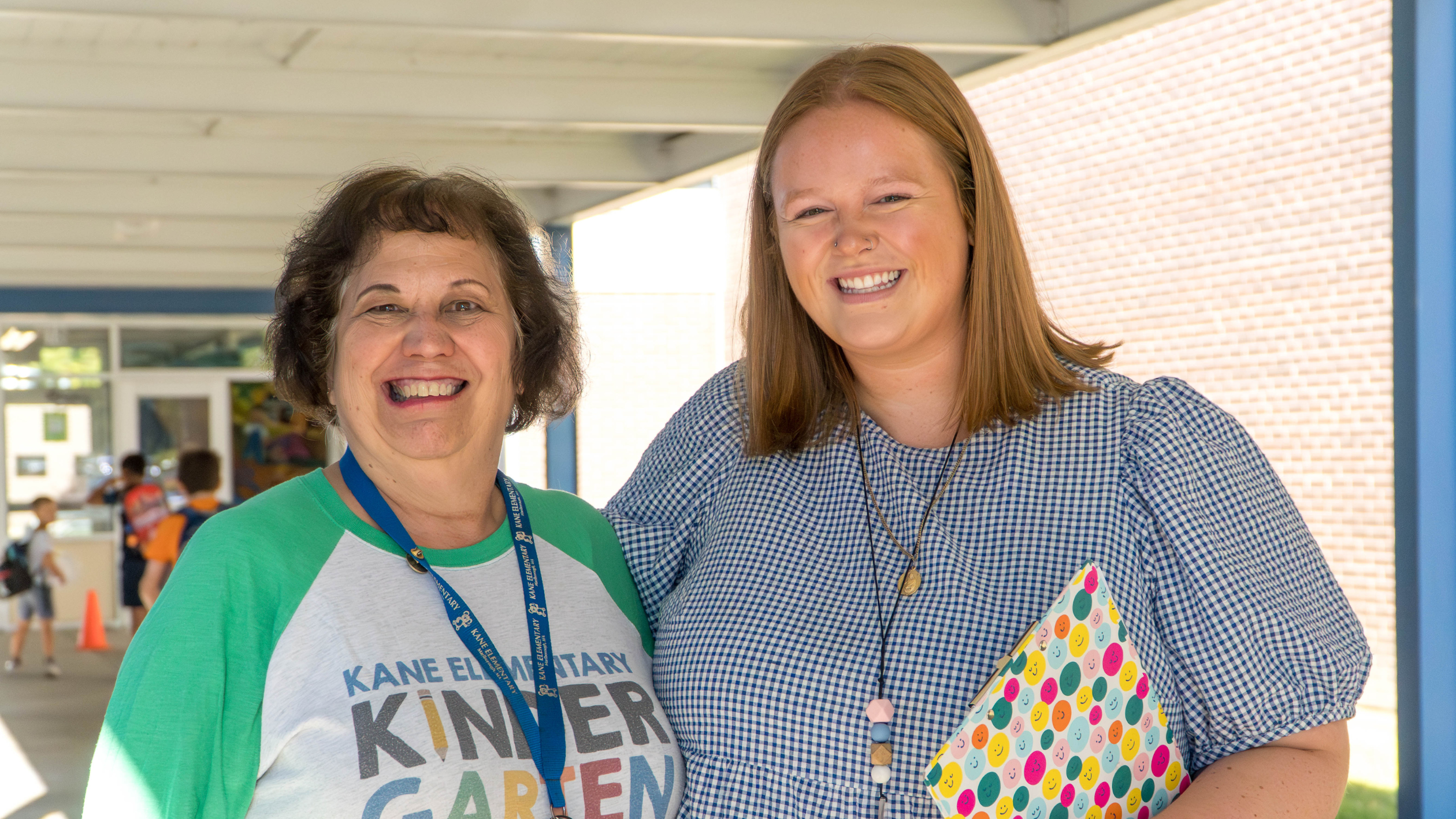Two women, one in a green shirt, one in a blue shirt, smile at the camera in a hallway.