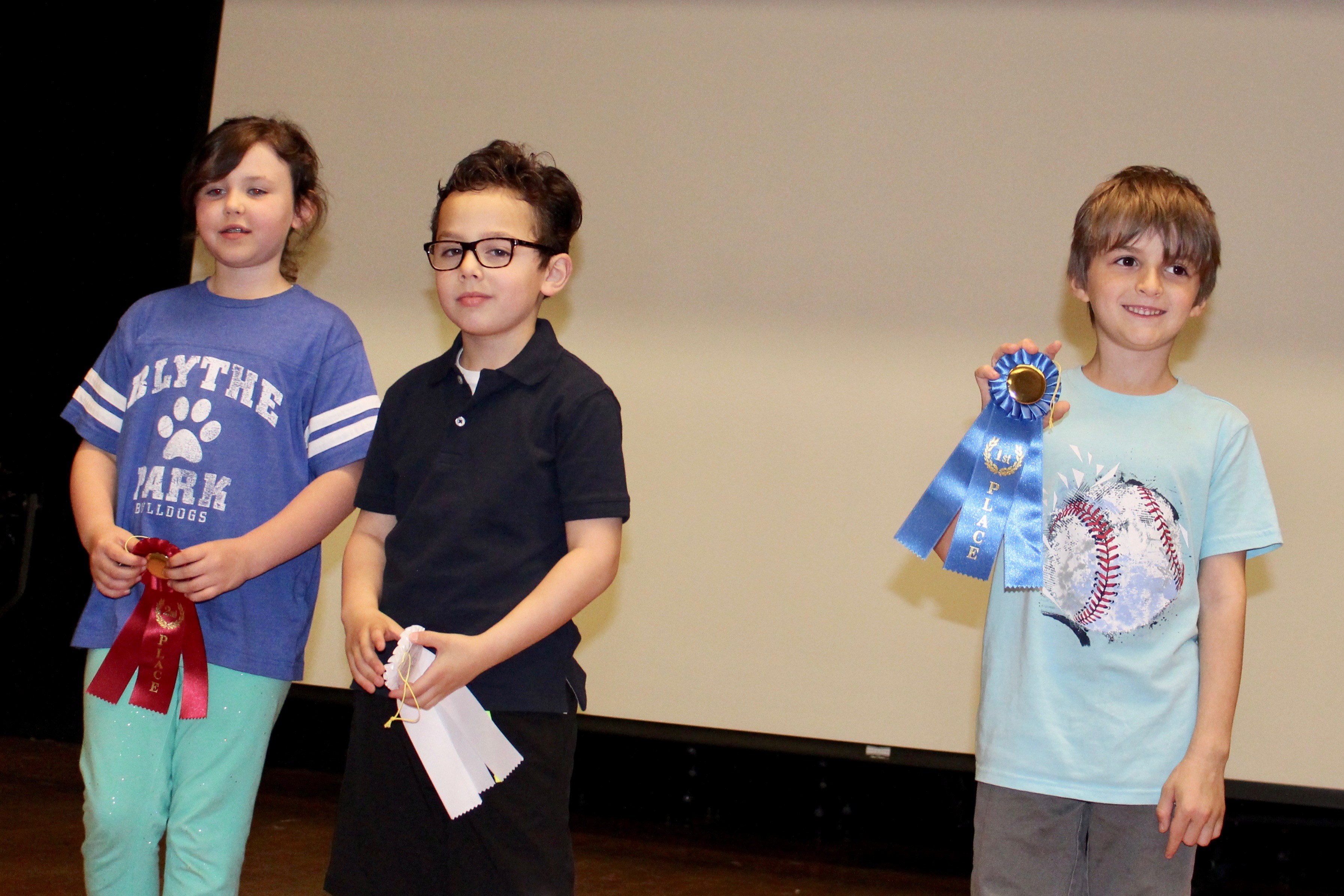 Three children proudly displaying their awards on a stage, celebrating their achievements.