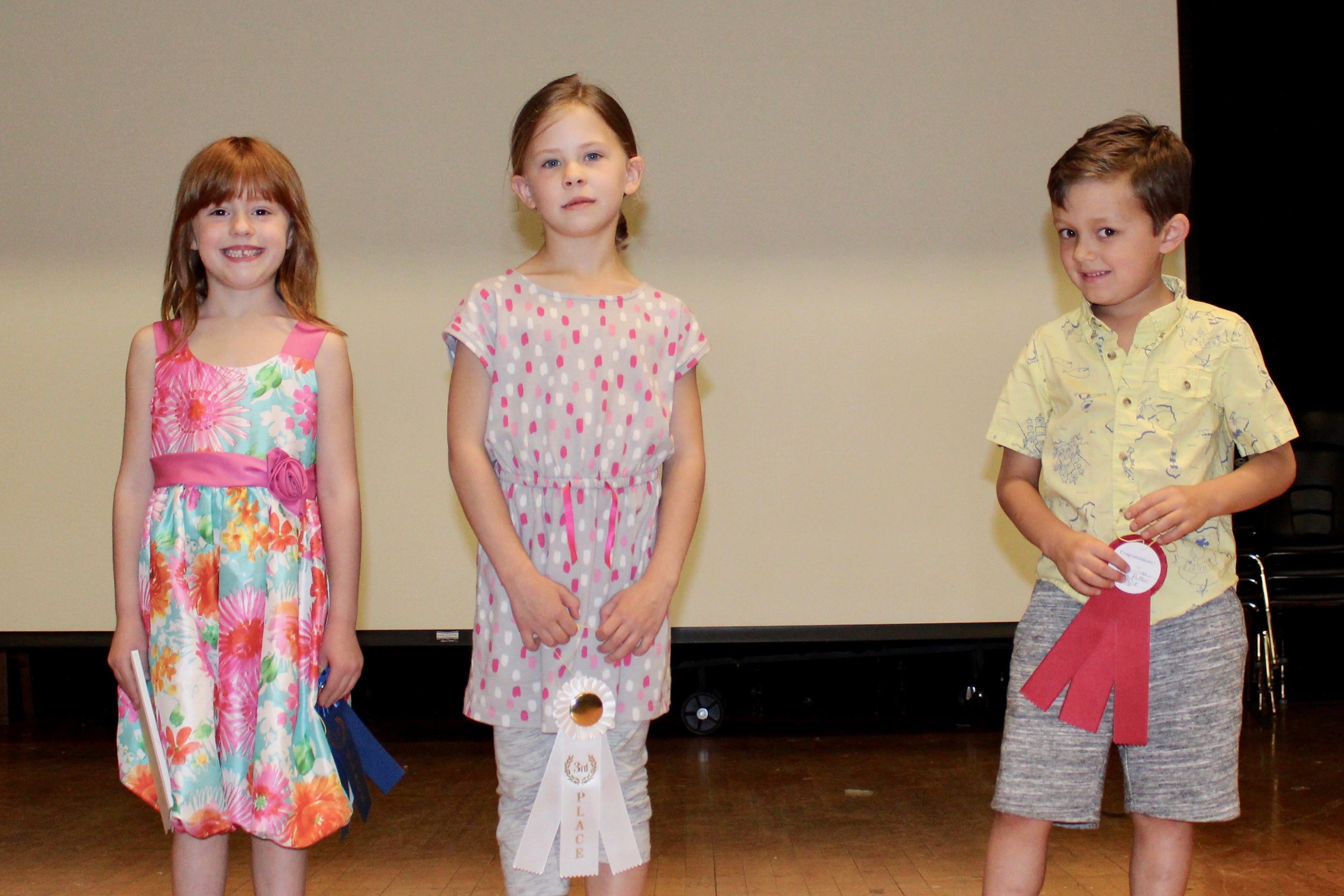 Three children on stage, proudly holding ribbons they won.