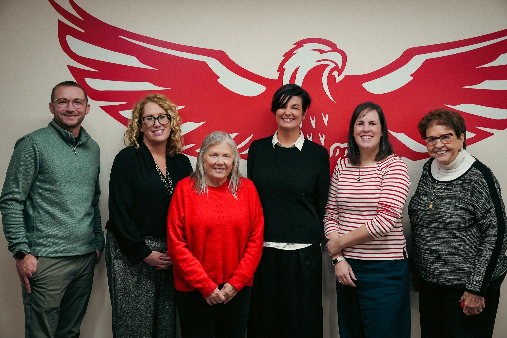A group of 5 people standing in front of a wall with a red eagle on it. From left to right: Man in a green quarter-zip, a woman with blonde hair and glasses, a shorter woman with a red sweater, a woman wearing a black sweater with a white collar, a woman in a red and white stripped shirt, a woman with brown hair and glasses.