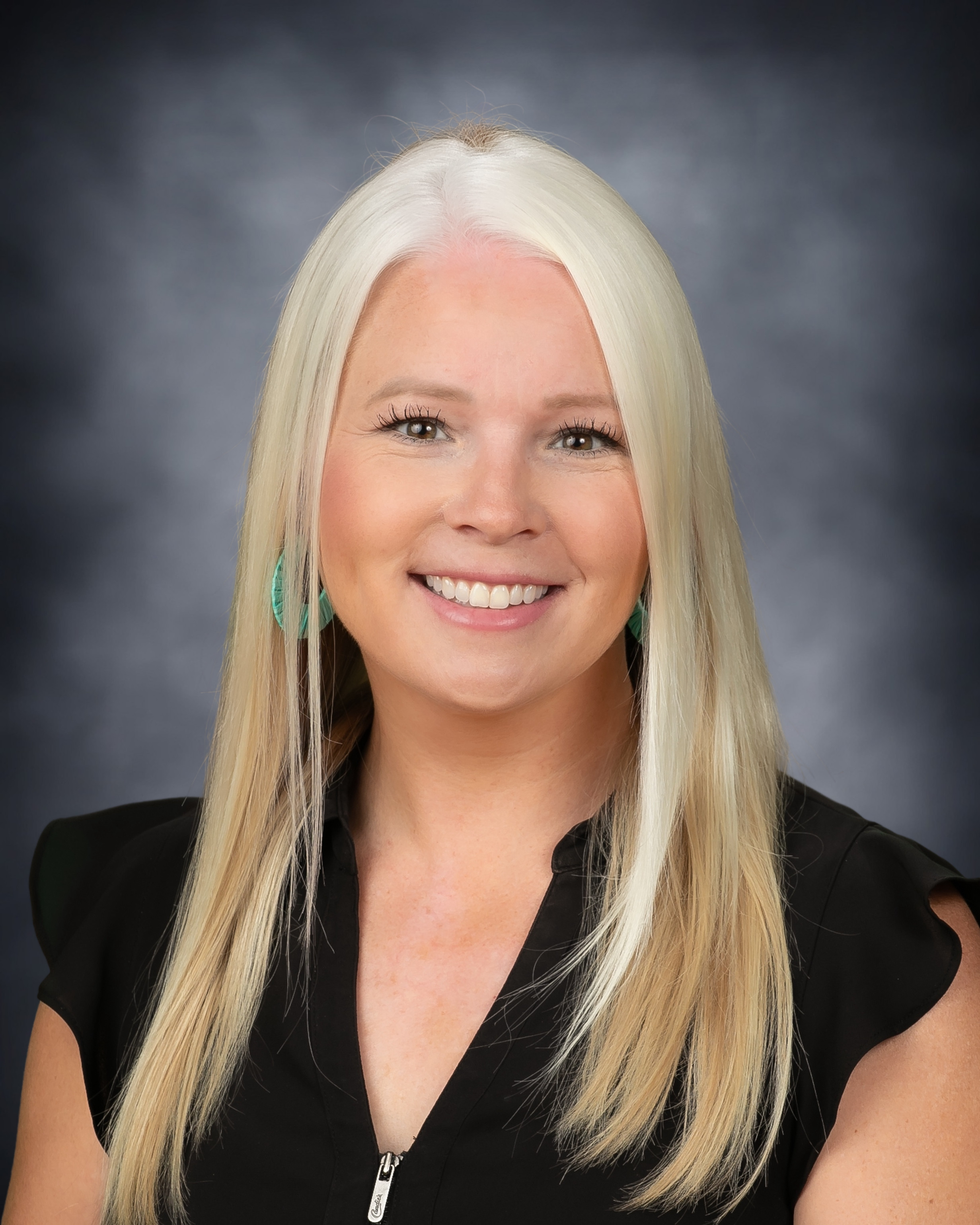 Professional headshot of a smiling woman with long, straight blonde hair wearing a black top and turquoise earrings against a gray studio background.