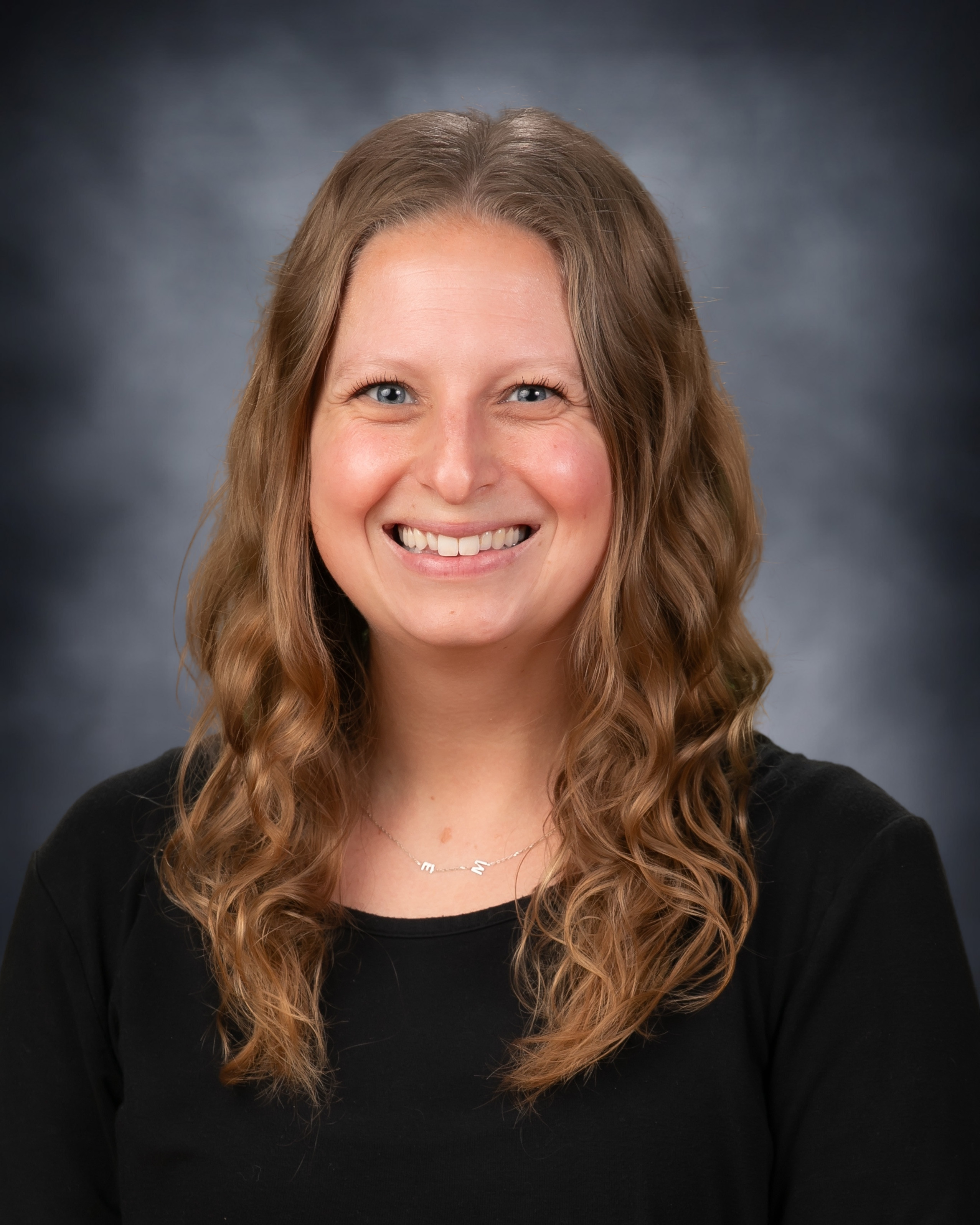 Professional headshot of a smiling woman with long, wavy brown hair wearing a black top and a delicate necklace against a gray studio background.