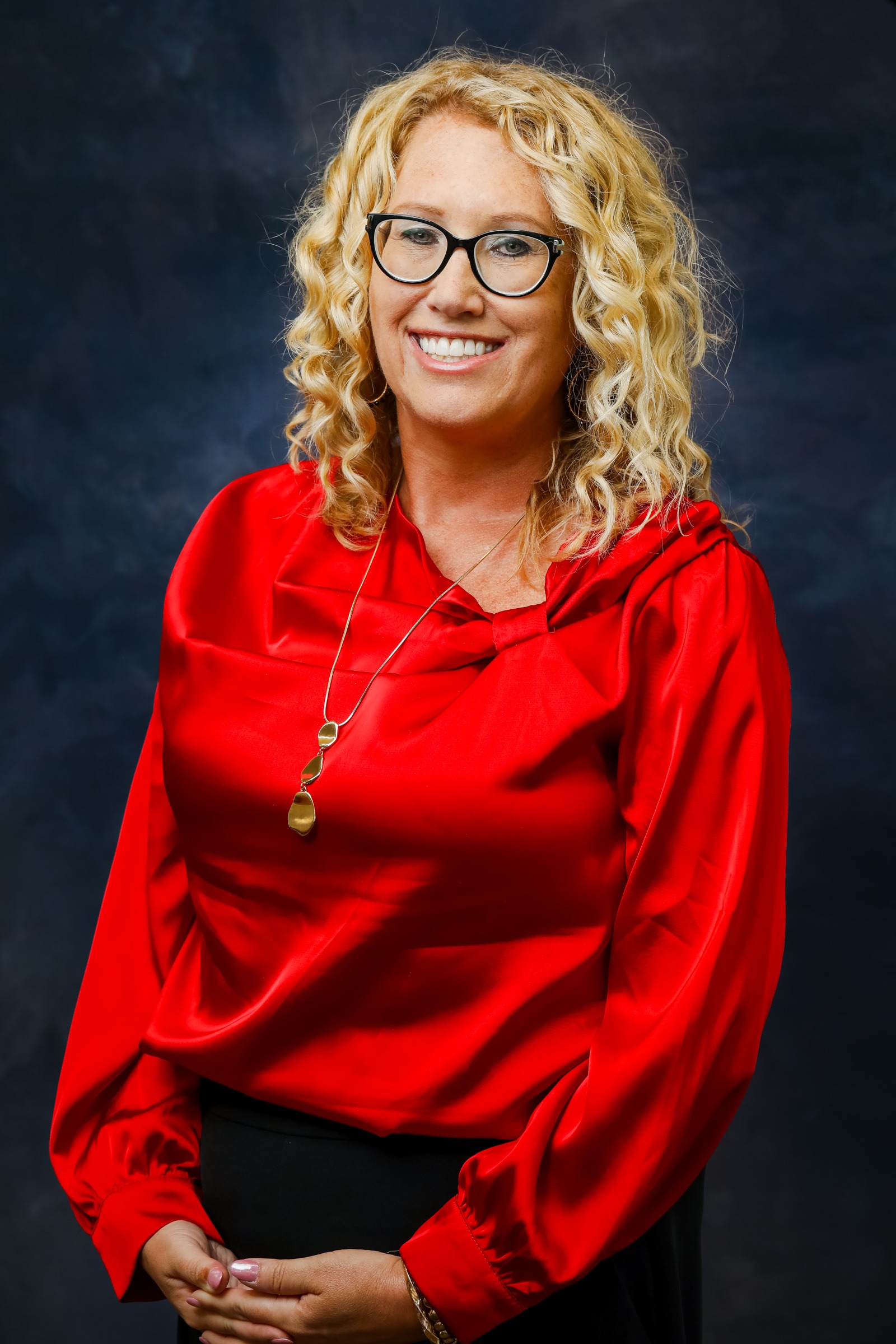 A woman with curly blonde hair and black glasses smiles for a professional portrait. She is wearing a bright red blouse and a gold necklace against a dark blue-gray studio background.