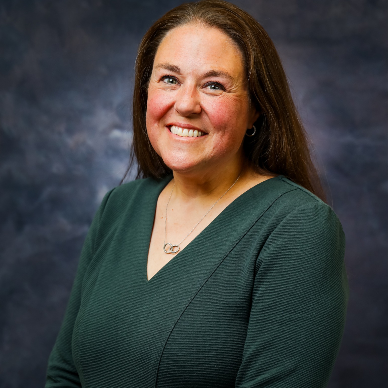 A brunette woman smiles for a professional portrait while wearing a dark green dress and silver necklace against a dark blue-gray studio background.