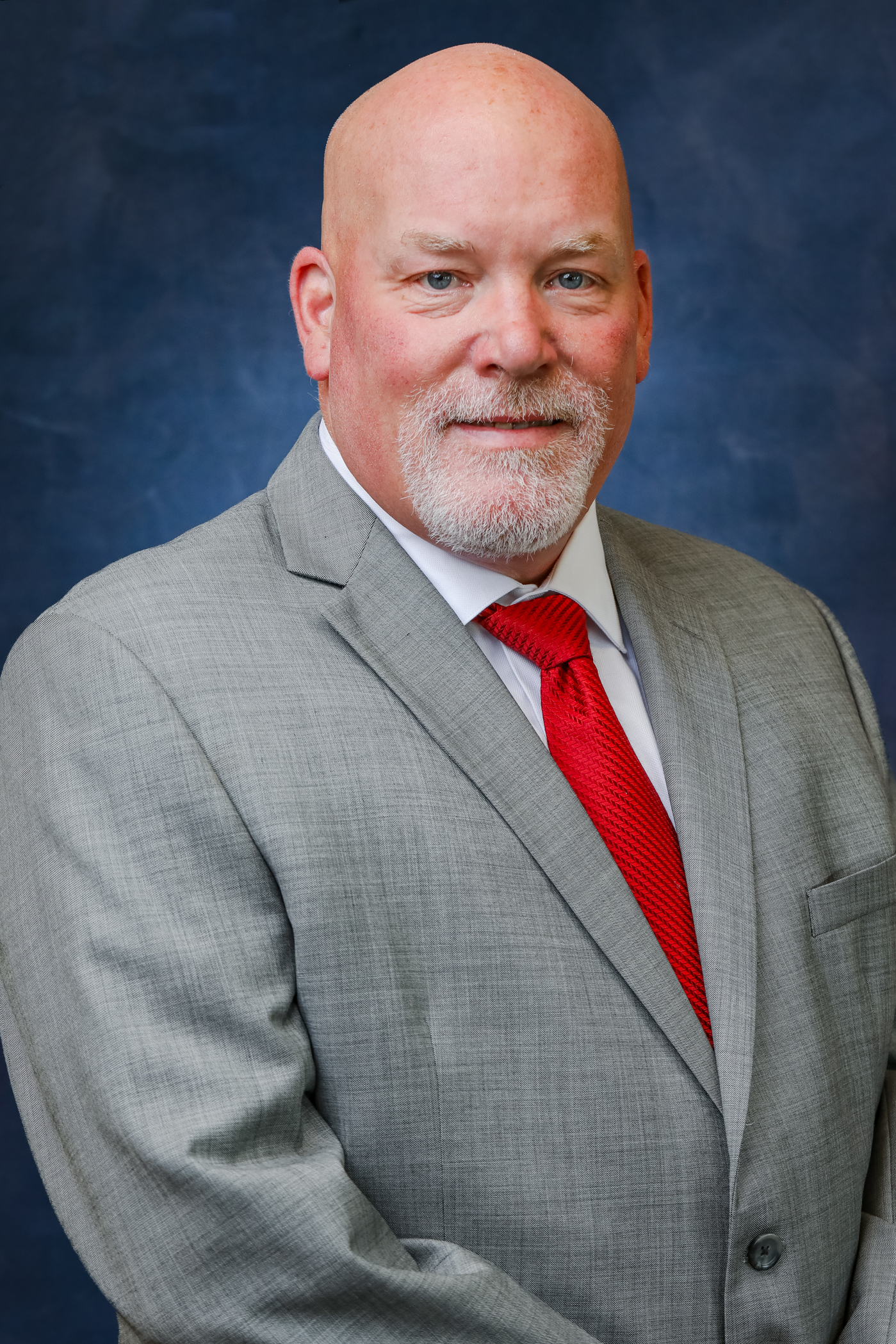 A bald man with a grey beard wearing a grey suit with a red tie