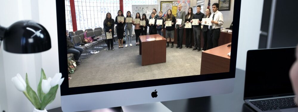 A group of people holding certificates displayed on an Apple computer monitor, with a desk lamp and flowers to the left and another laptop to the right