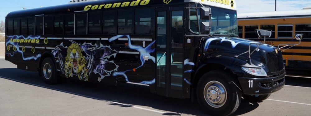 Two school buses parked side-by-side in a school bus parking lot. The bus on the left is black with white lettering that says "Coronado" and "Leopards." The bus on the right is a traditional yellow school bus.