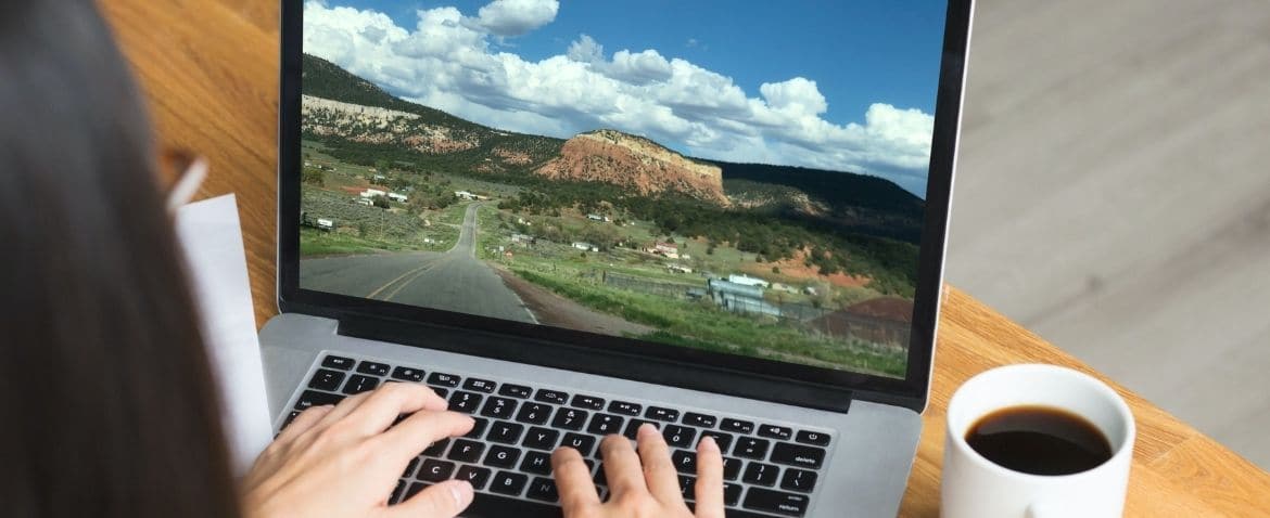 A person is typing on a laptop that displays a vivid image of a scenic landscape, with a cup of coffee beside it.