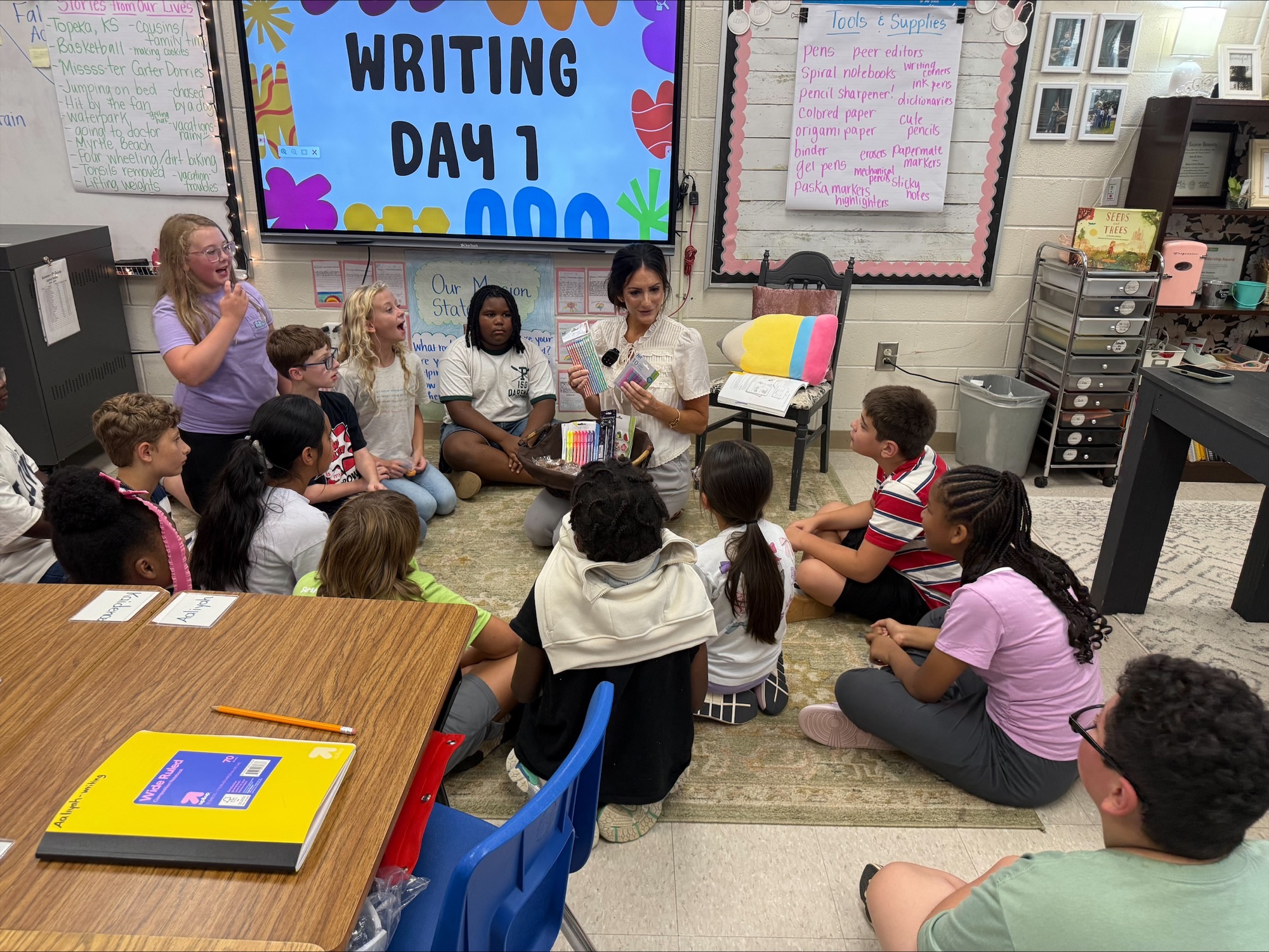 A classroom with children seated in a circle around an adult, listening to a story on a screen.