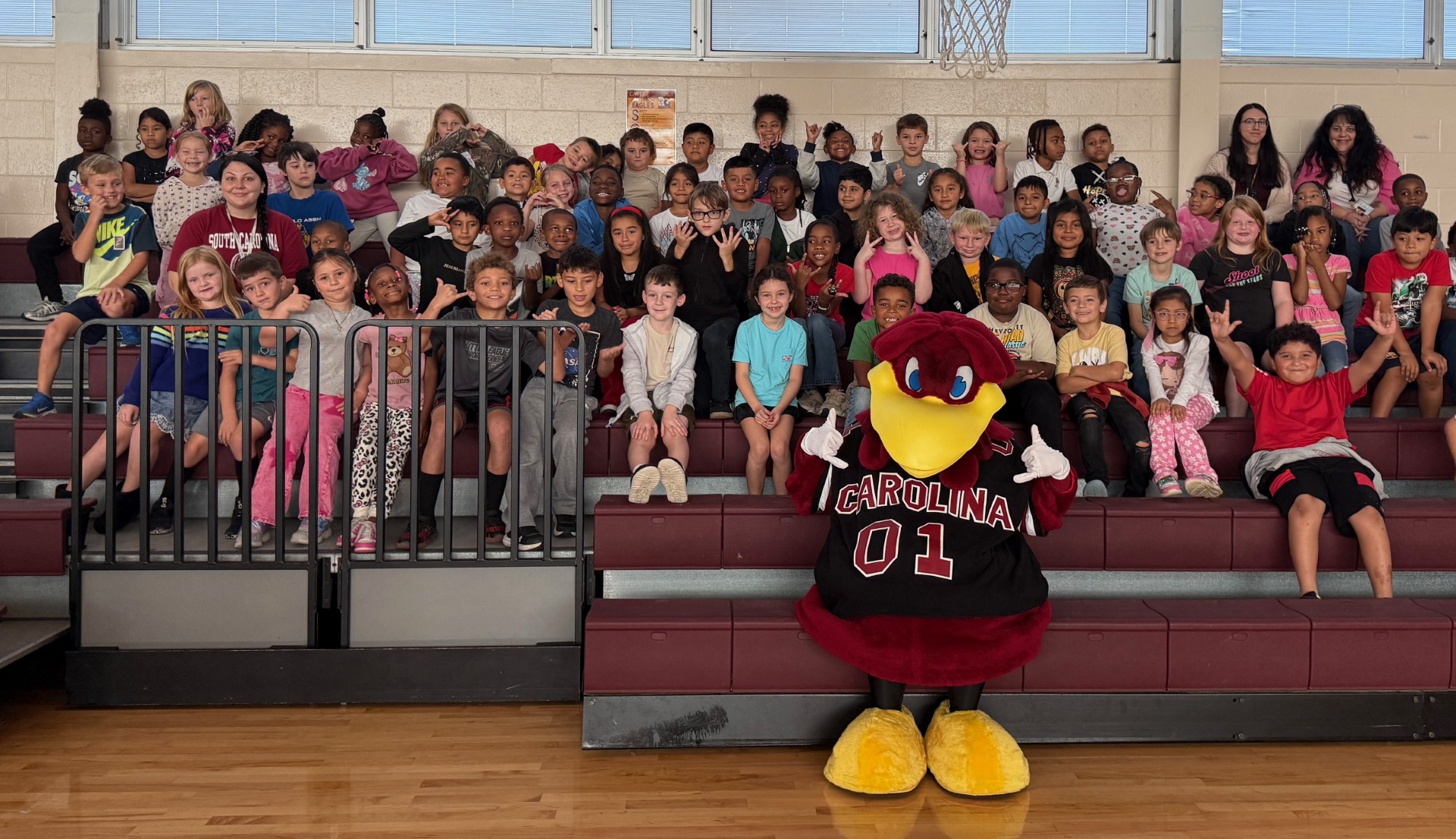 Second grade students are seated on bleachers, while the USC mascot, Cocky, poses in front of them.