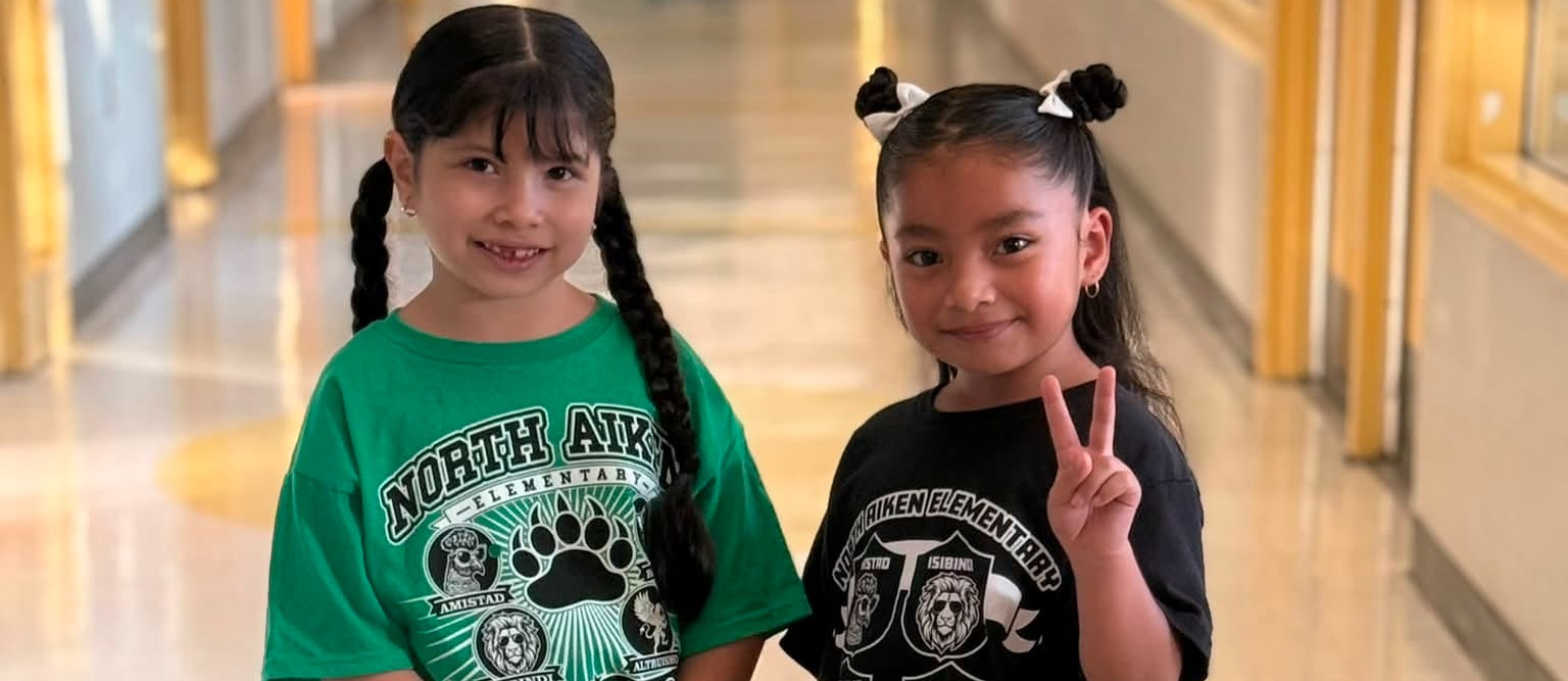 Two young girls, one in green, one in black, stand side by side in a hallway, making peace signs.