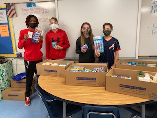 This is a image of 4 student standing around a large desk, wearing a mask, and holding up toiletry items 