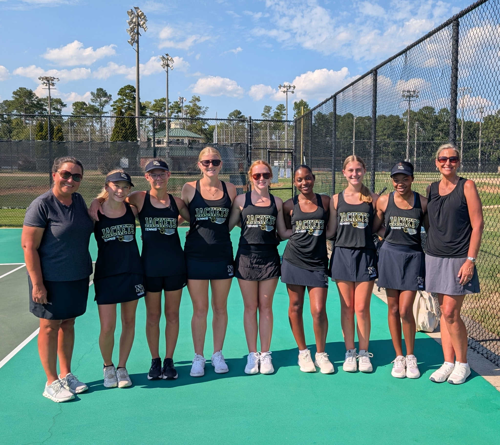Nine individuals stand in a row on a tennis court with a black fence; six wear identical black outfits with a logo.
