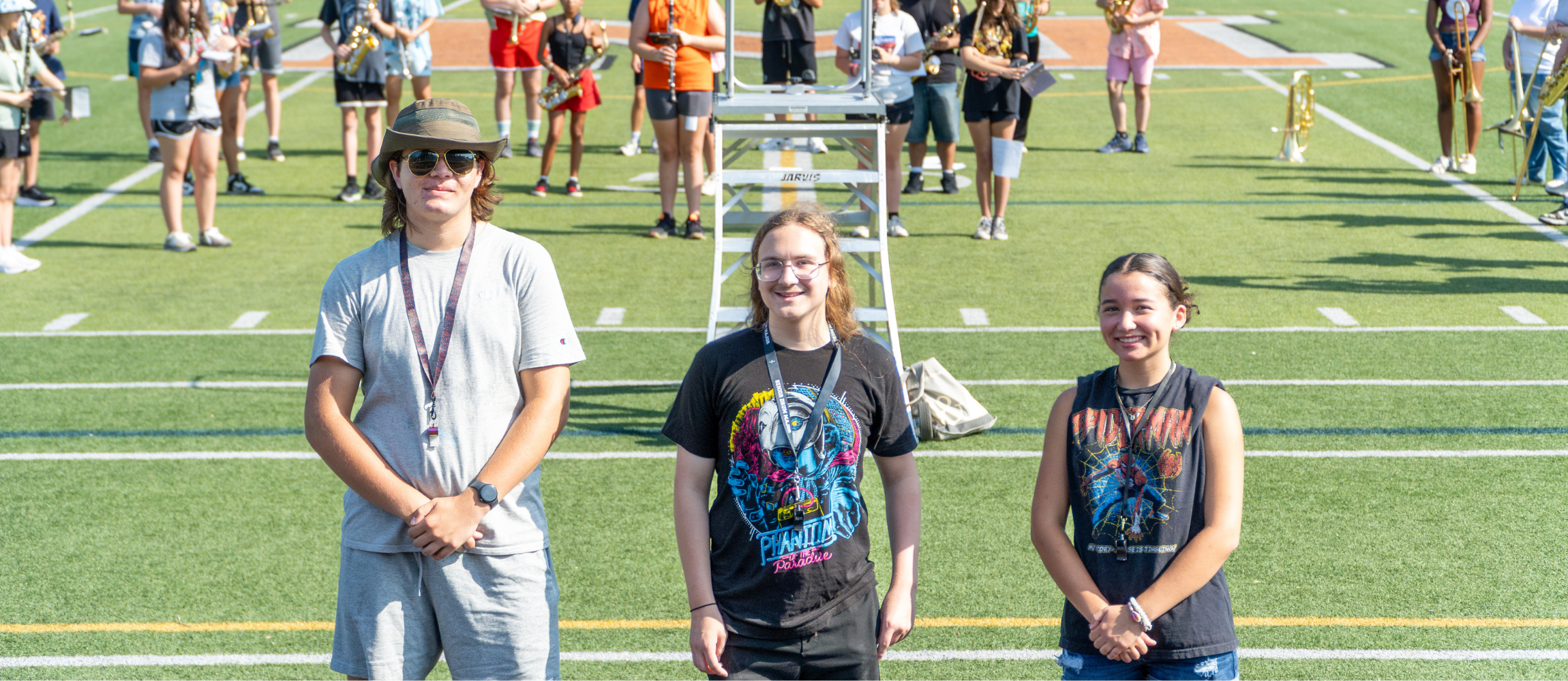 The three drum majors for the 2025-2026 school year pose for a group photo on Noble Field. The rest of the band stands behind them.