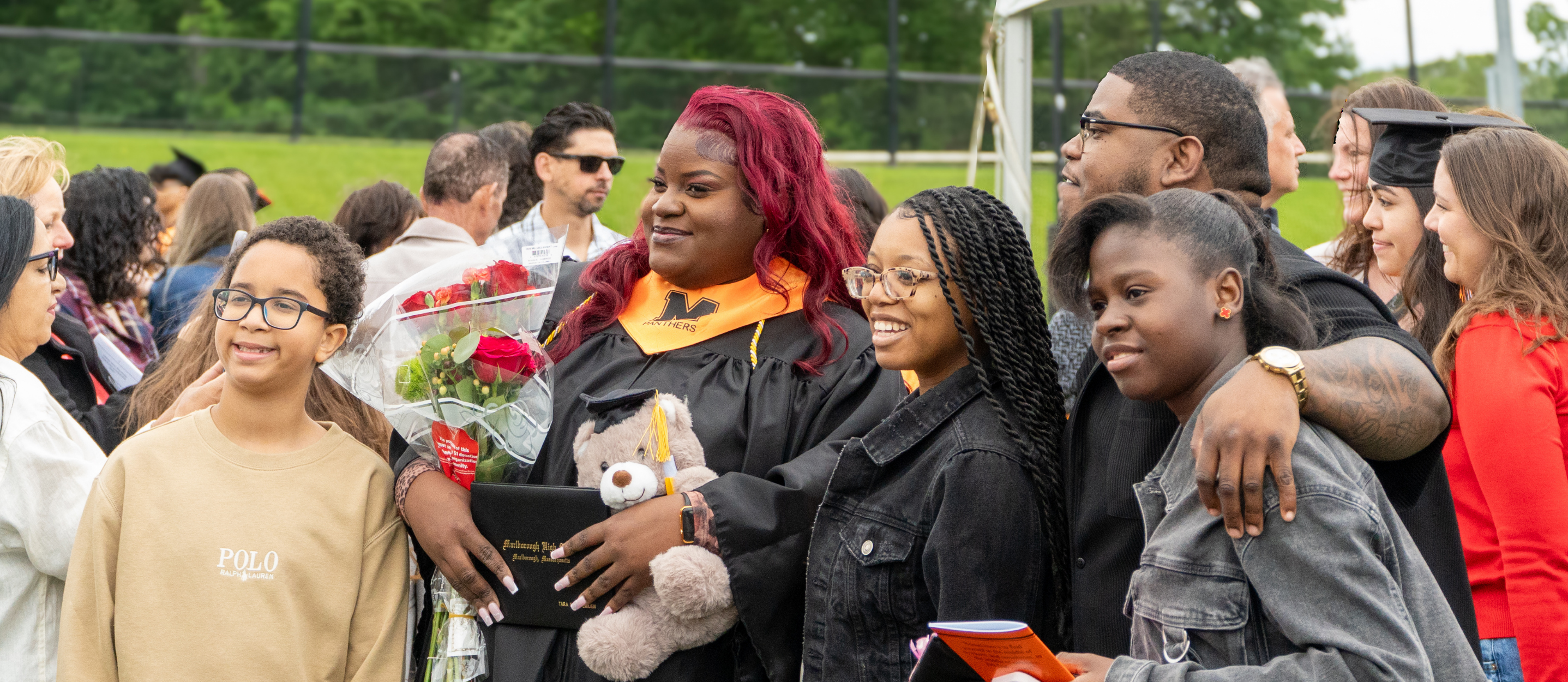 A group of people, some in graduation attire, stands in an outdoor area with a green lawn.