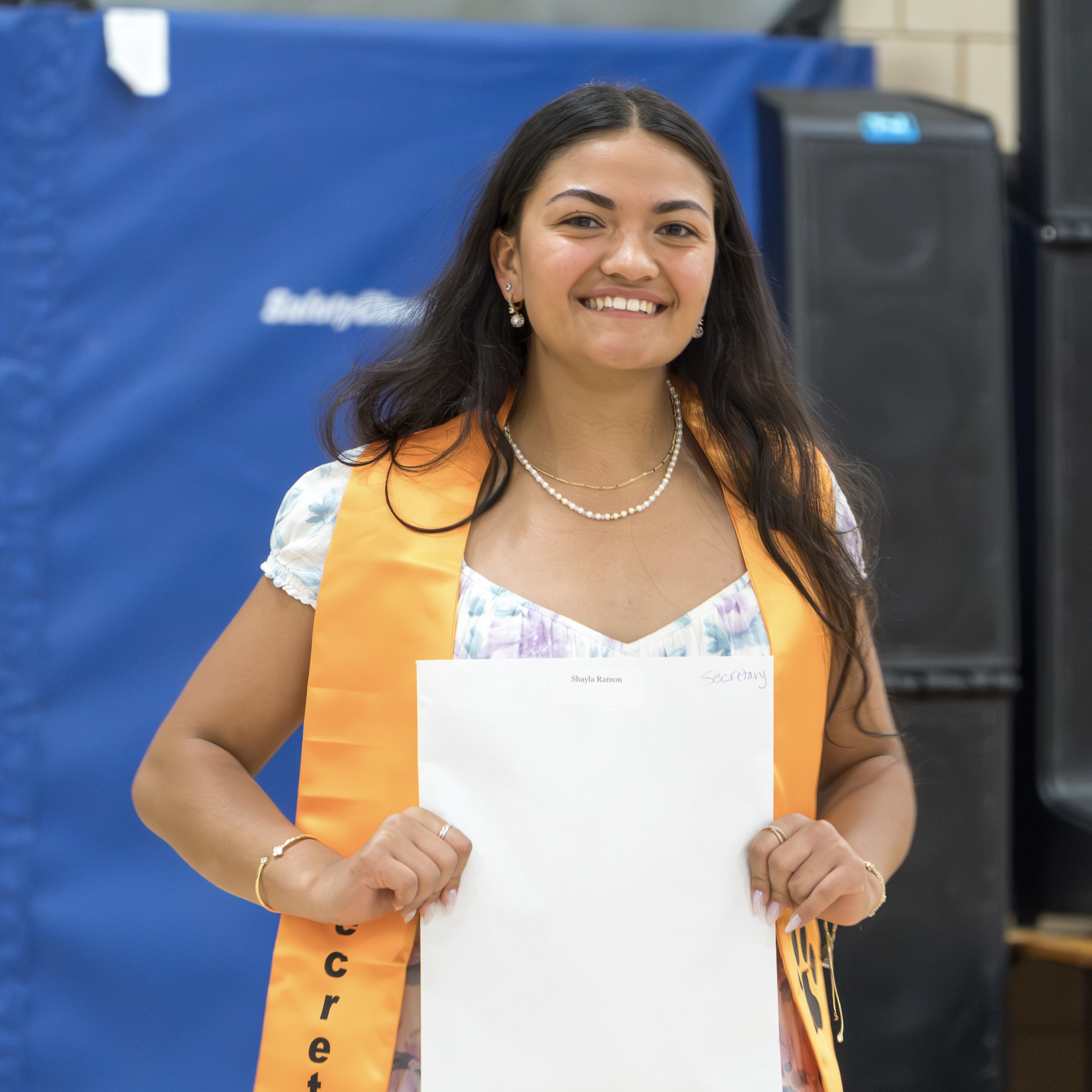 A woman in a floral top and orange sash smiles and holds a paper in front of a blue wall.