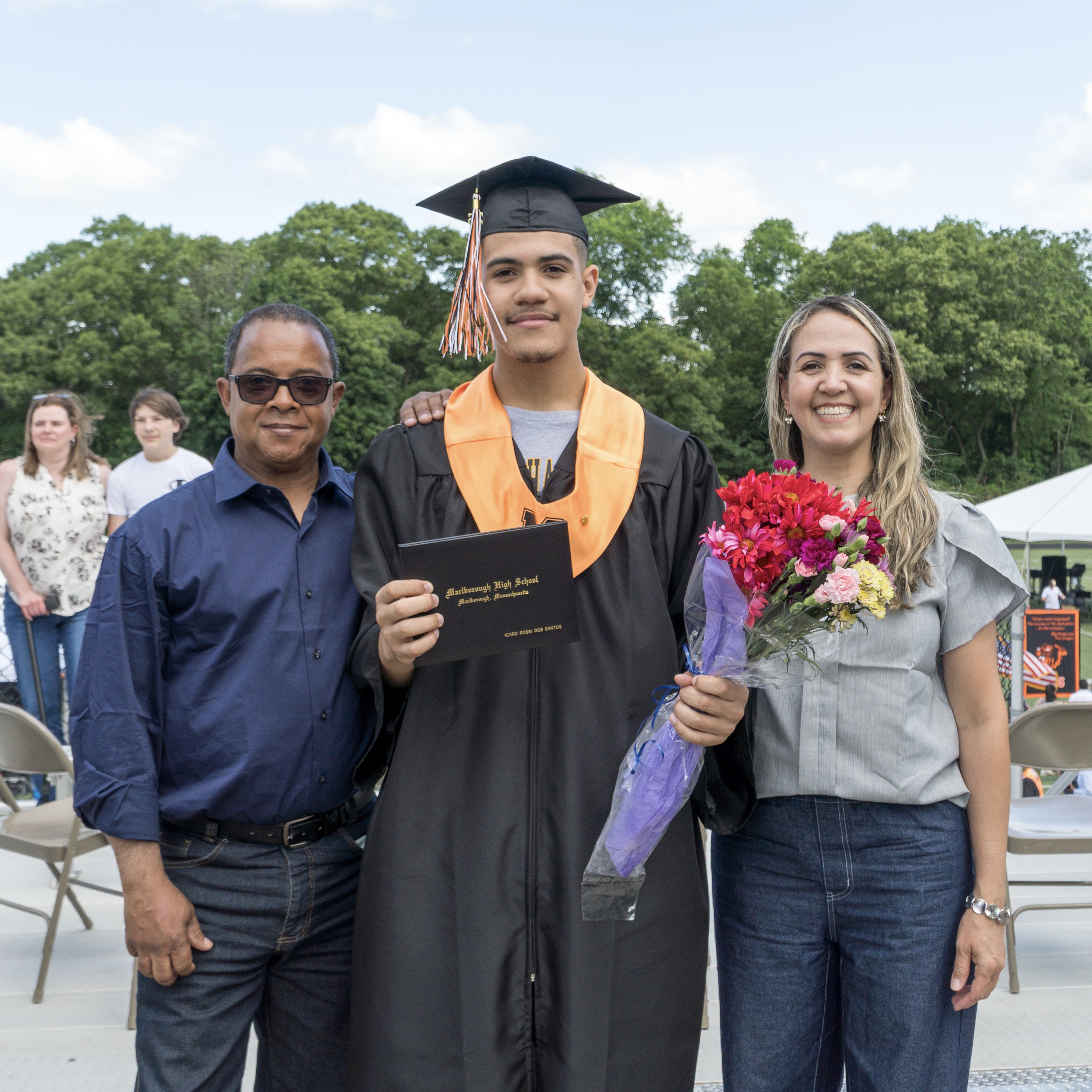 A man in a cap and gown holds a certificate next to a woman with flowers. Behind them, a man in a blue shirt stands. In the background, people sit in chairs.