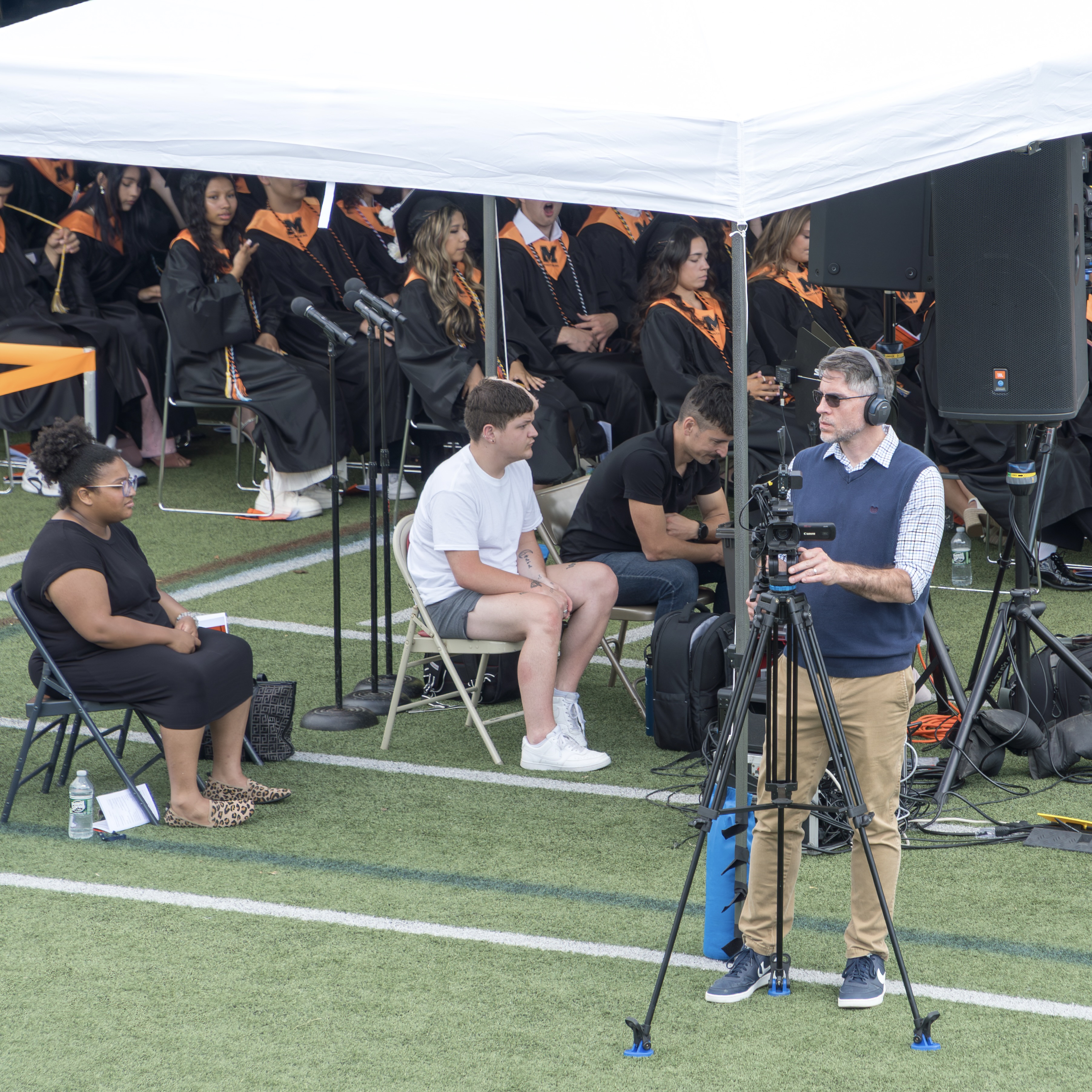 People seated on chairs under a white tent, with a person standing with a camera on a tripod.