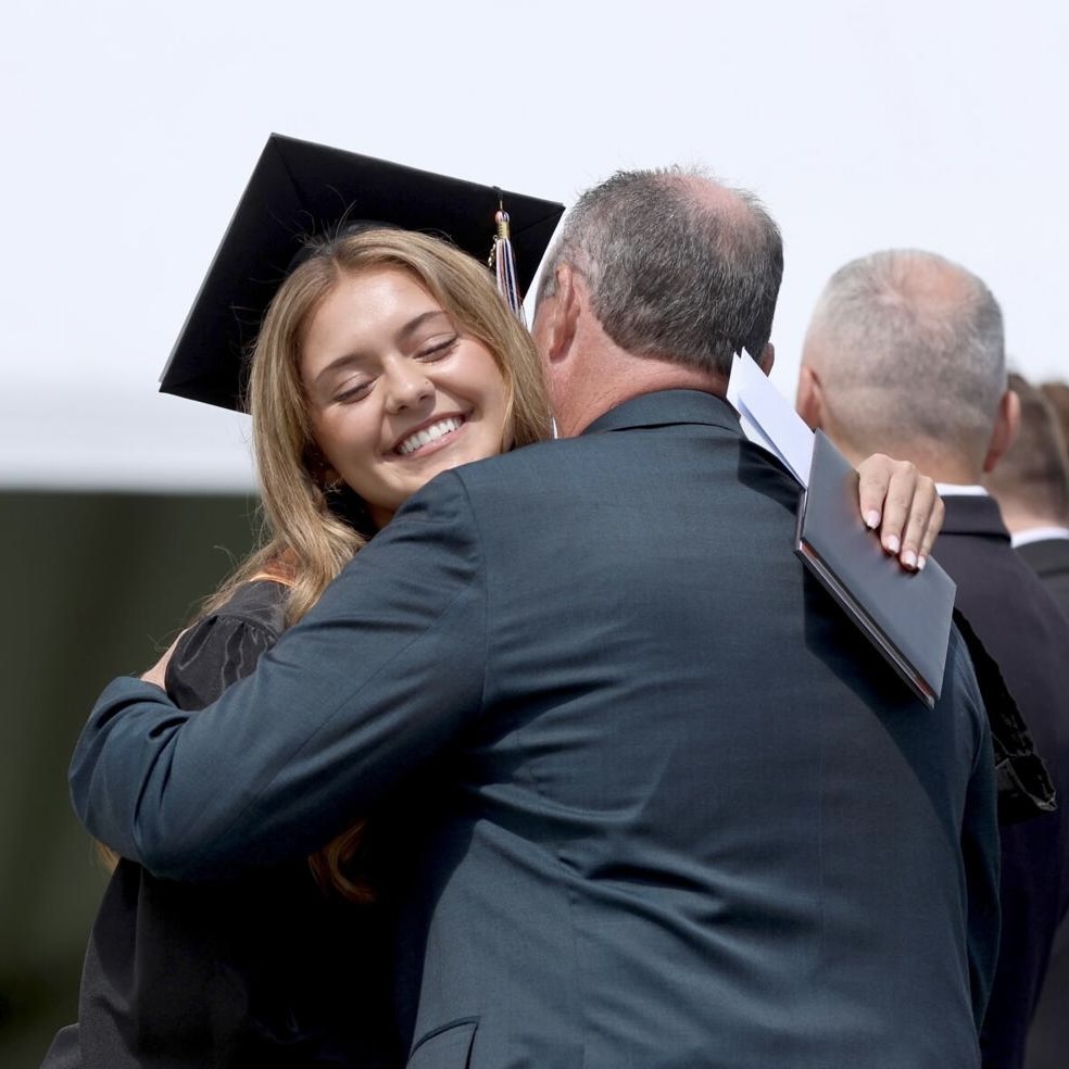 A graduate in cap and gown hugs an older man in a suit during an outdoor ceremony.