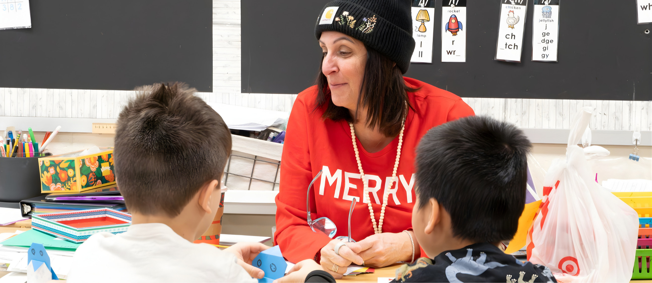 A Jaworek teacher sits at a desk with two students, one looking at her and the other holding a blue object.