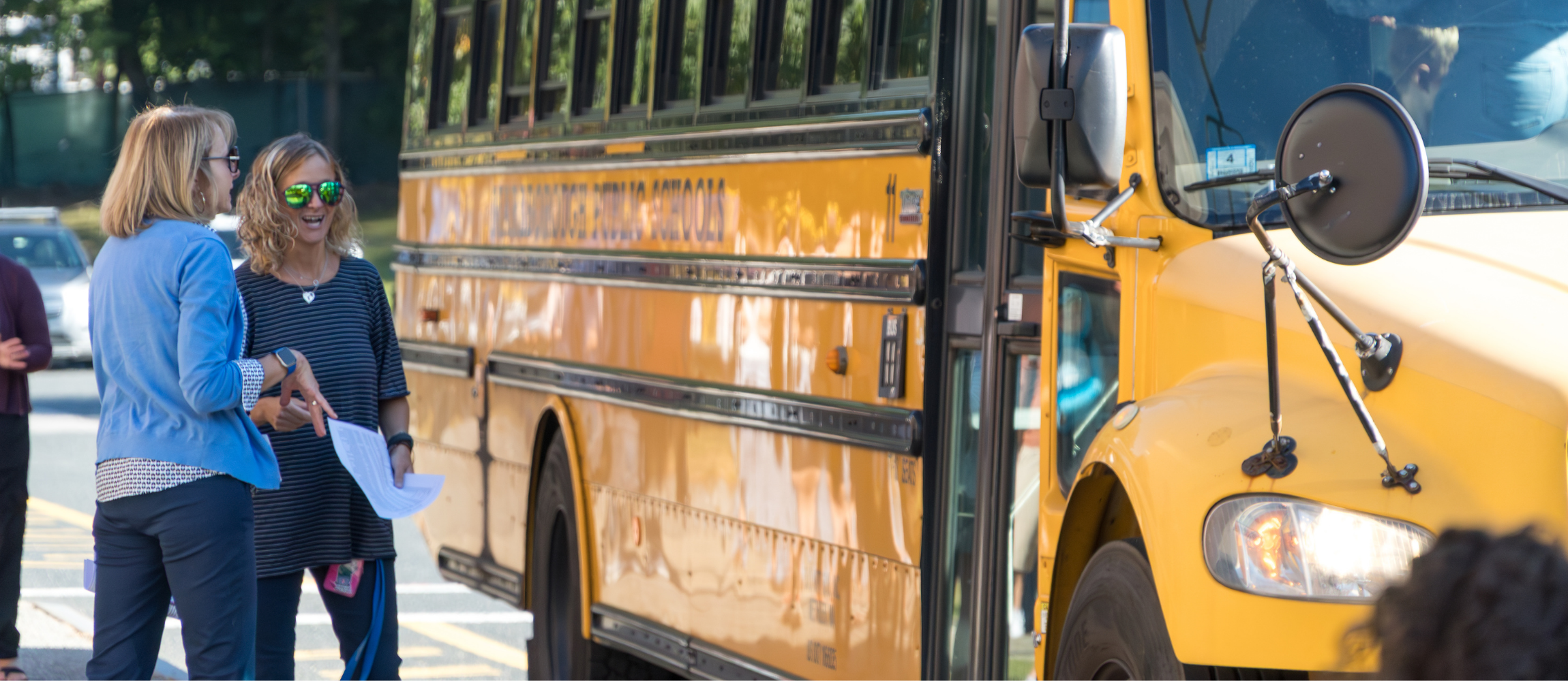 Kane School Principal KP and another staff members smile while standing next to a yellow bus.
