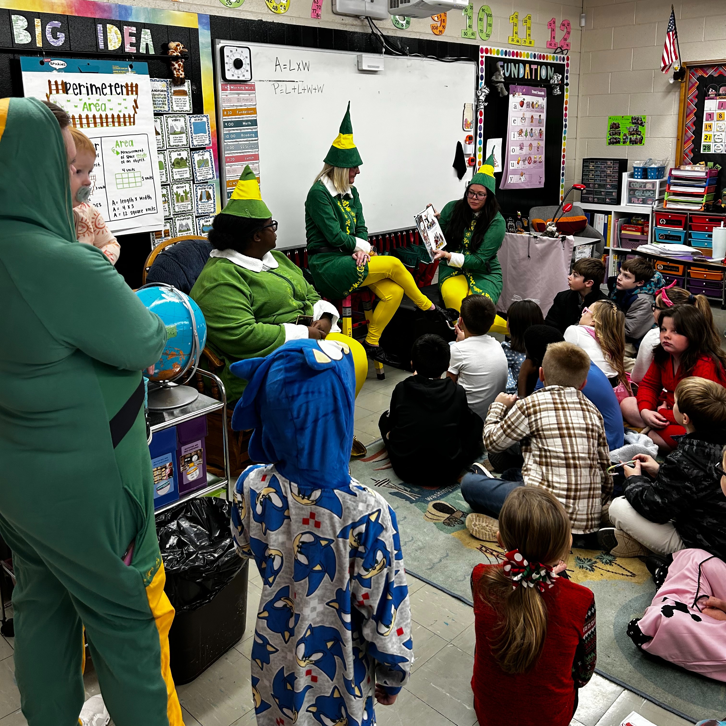 Three staff members dressed as elves sit at the front of an elementary classroom reading a picture book aloud to a group of students seated on the carpet. The classroom walls display colorful math posters, number decorations, and a whiteboard, while students listen attentively during the festive read-aloud activity.