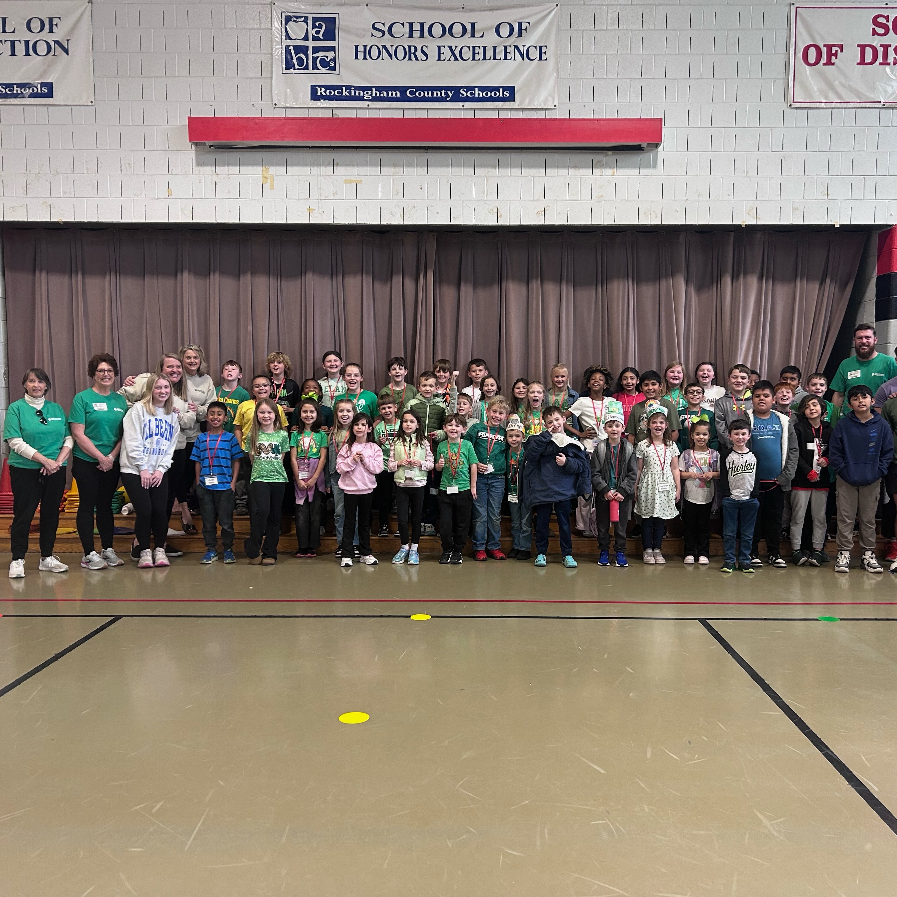A large group of elementary students and staff stand together on a stage in a school gymnasium, many wearing green shirts and medals on lanyards, posing for a group photo beneath banners reading “School of Distinction” and “School of Honors Excellence.”