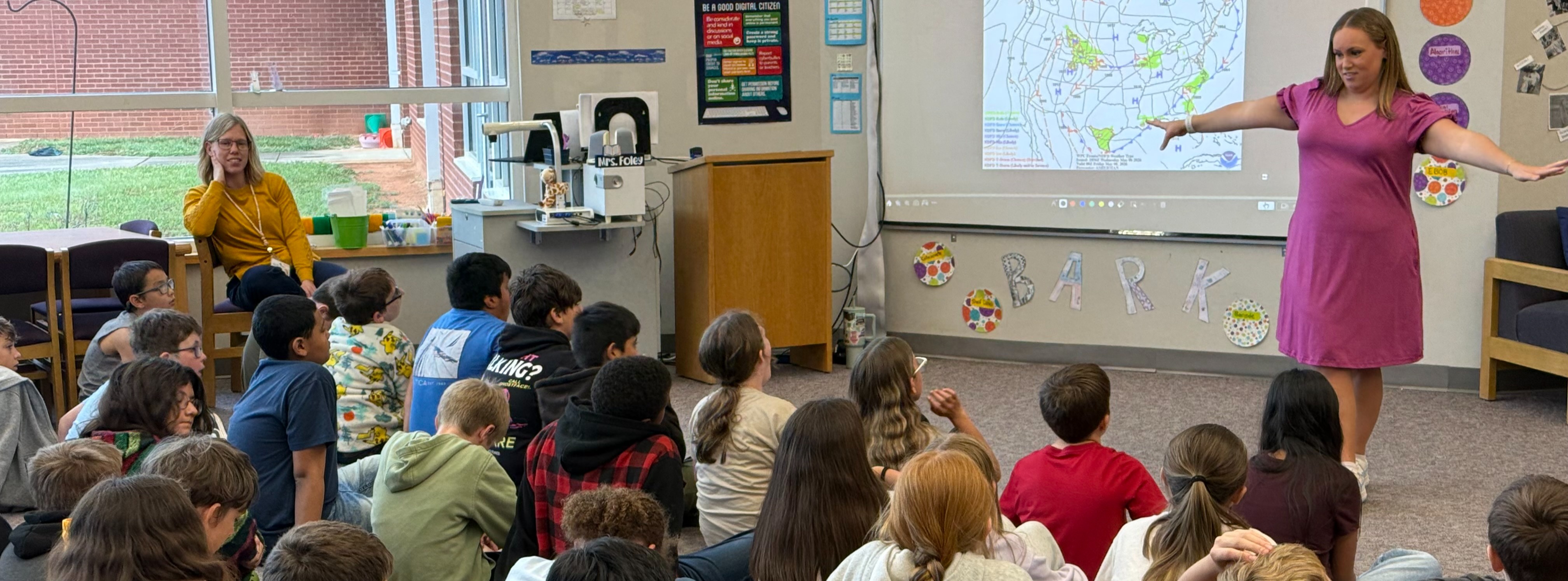 woman standing in front  of children on the floor showing a weather map on projector screen