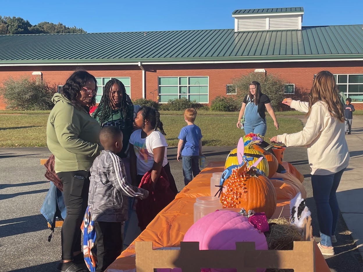 The image shows families gathered outside of a school during a parent night. Grade level teachers decorated pumpkins, and the pumpkins are displayed on a table. The families are viewing the pumpkins to vote for their favorite one.