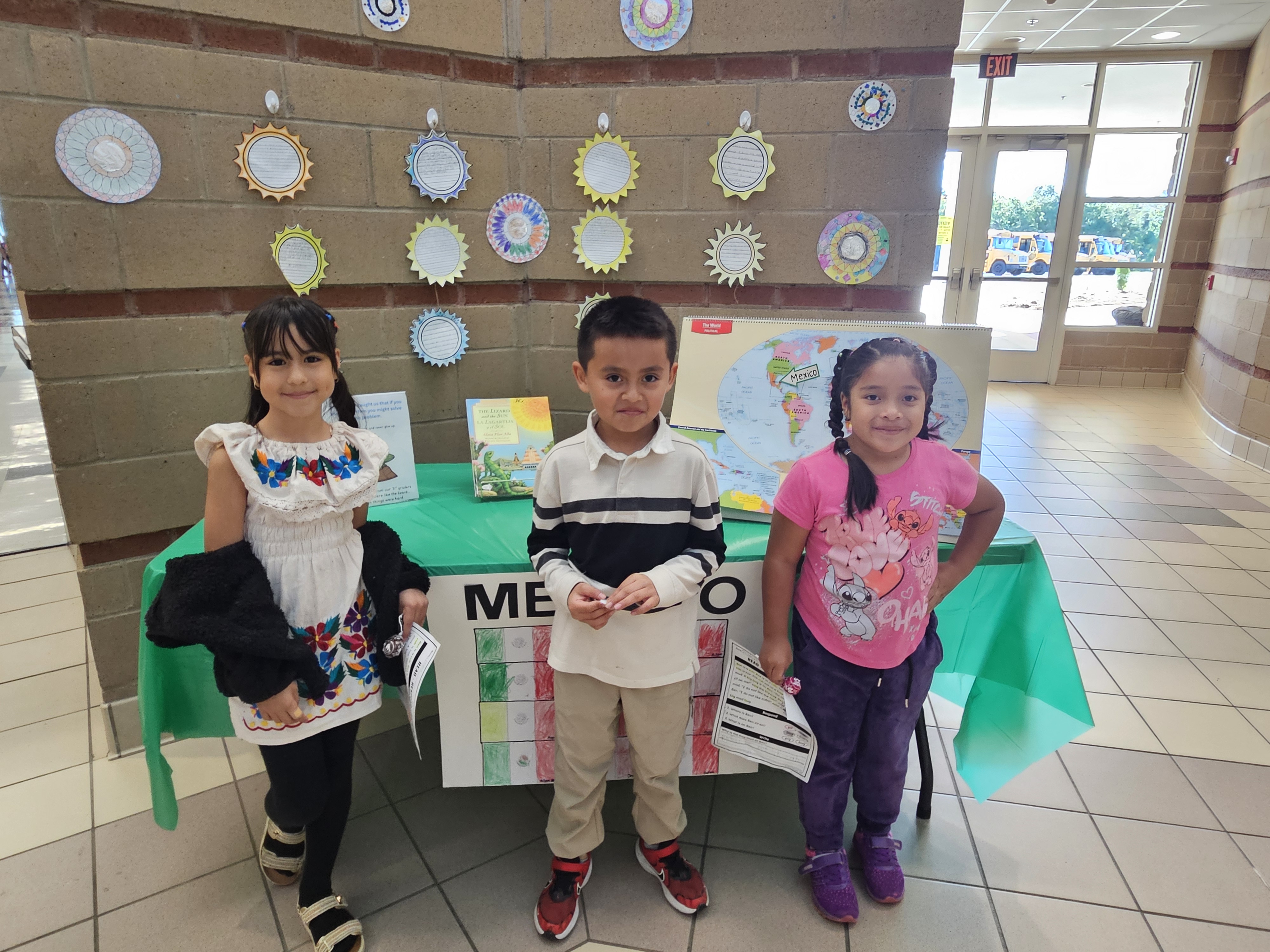 3 Students standing in front of a table with information about Mexico during a Hispanic Heritage program
