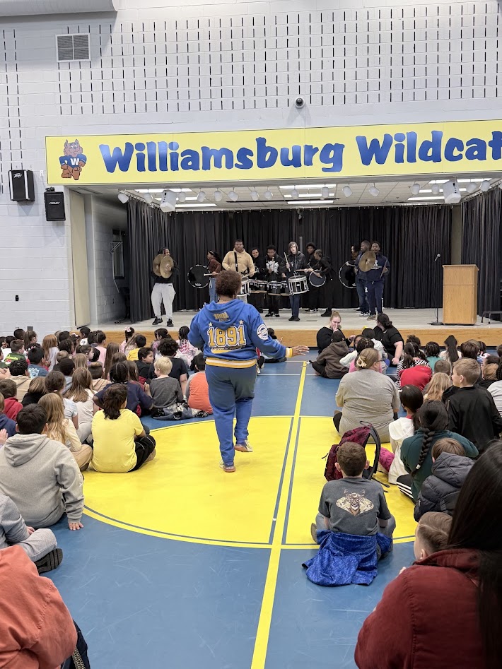 Image shows the guidance counselor leading students during a Black History Month assembly. Students are seated on the floor and the high school marching band is performing on stage.