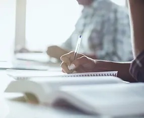 student writing in a notebook with an open textbook on desk