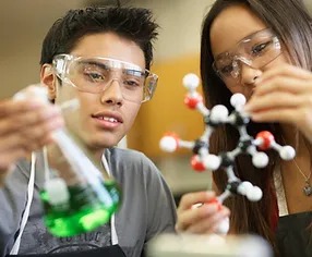Students wearing protective glasses in a Science Class using a beaker with green chemicals and an atom model