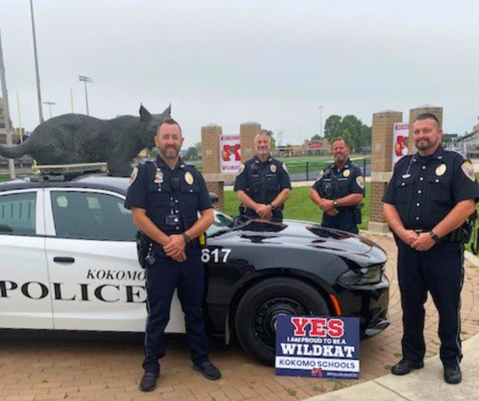 Student Resource Officers smiling kindly in front of police car with football field in background