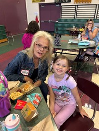 A proud woman poses with a young girl at a school event, both seated at a table with snacks and drinks.
