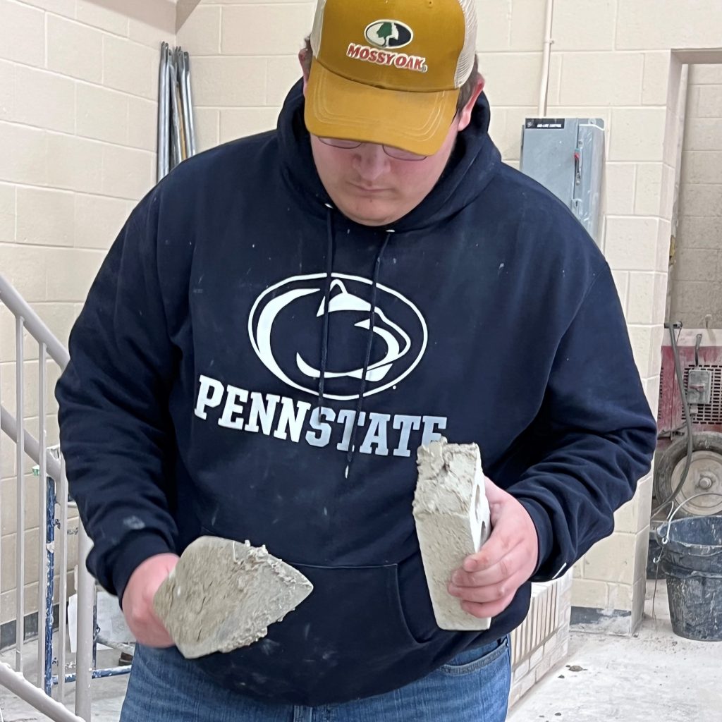 A person clad in a Penn State hoodie and cap is holding two large pieces of broken concrete