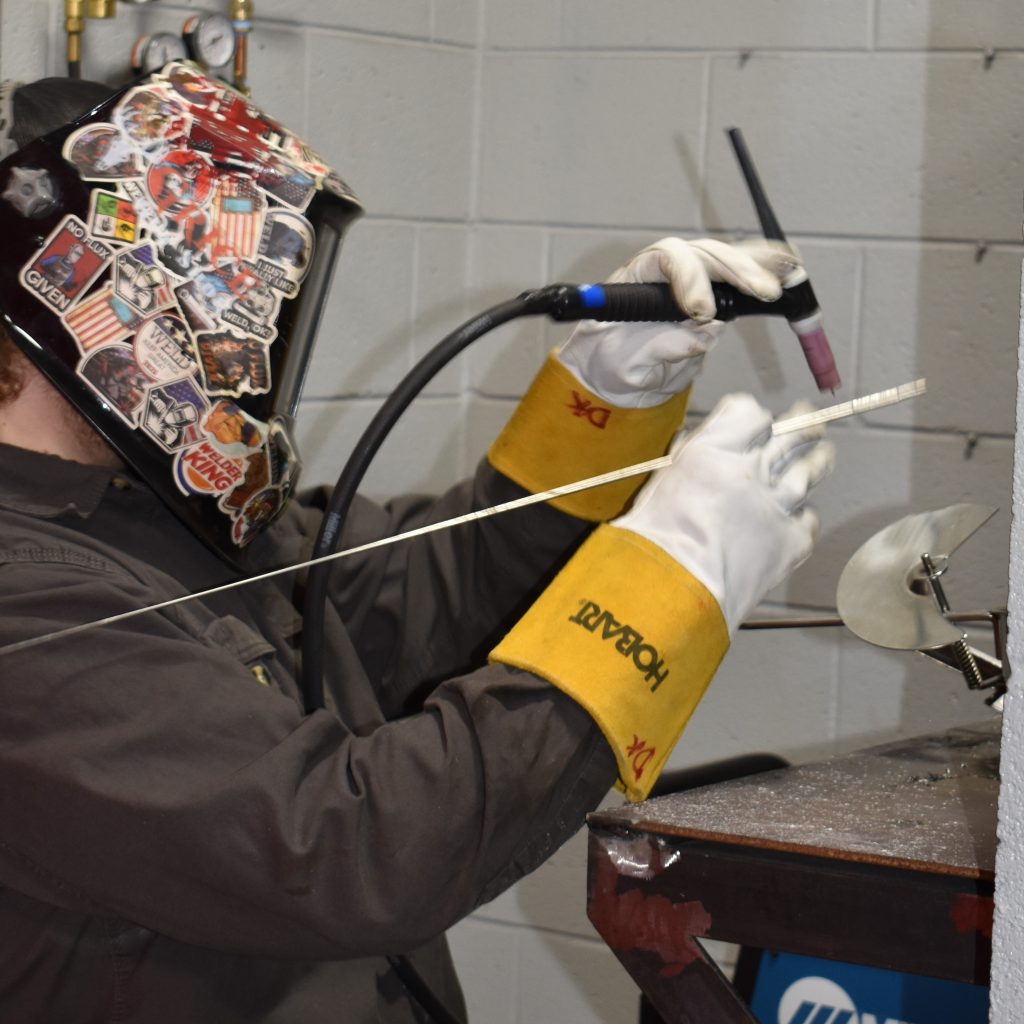 A person clad in protective gear, including a helmet adorned with various stickers, is engaged in the skilled task of welding