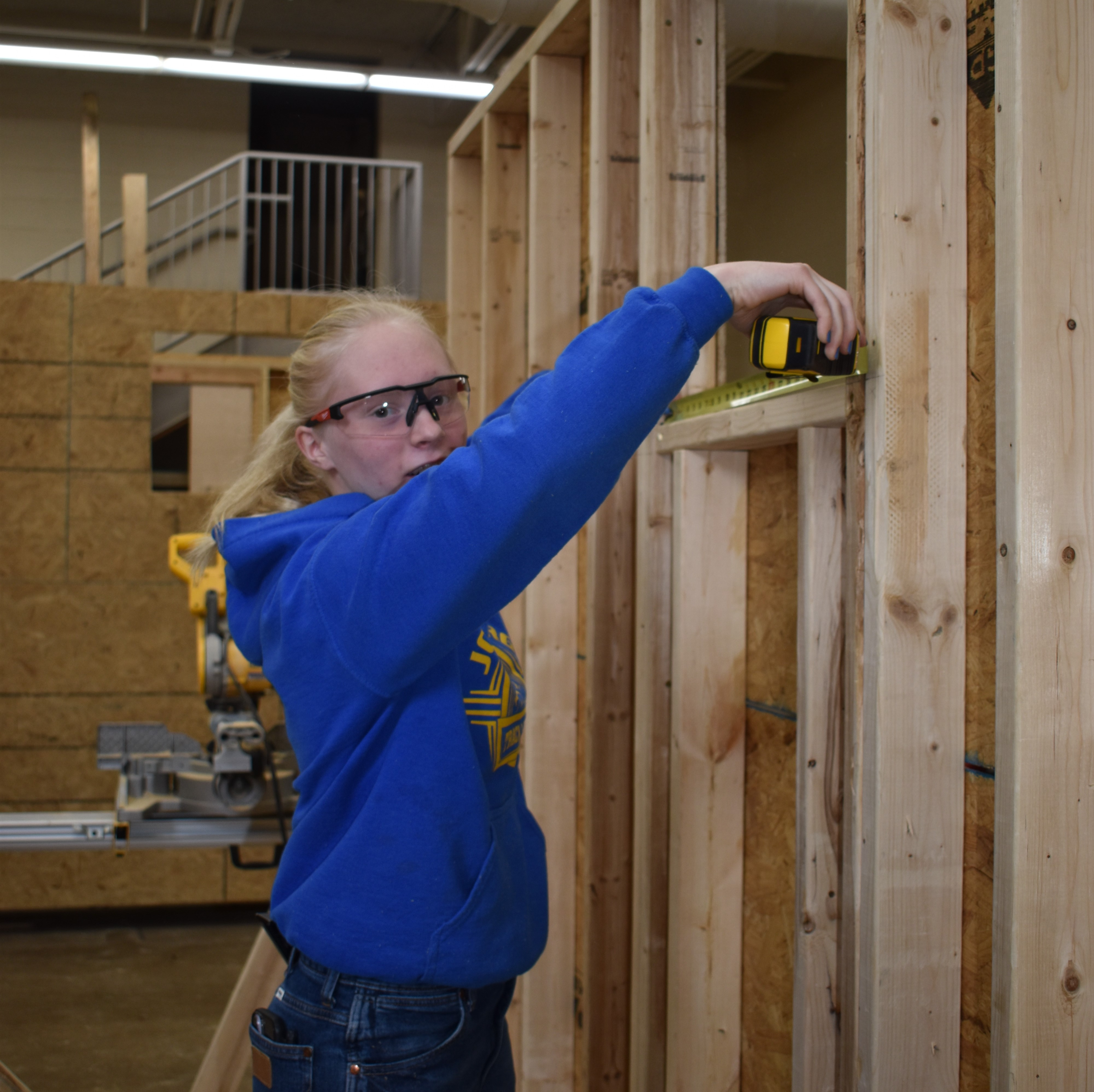 A person in a workshop, using a measuring tape on a piece of wood placed on a table, surrounded by various woodworking tools and materials.