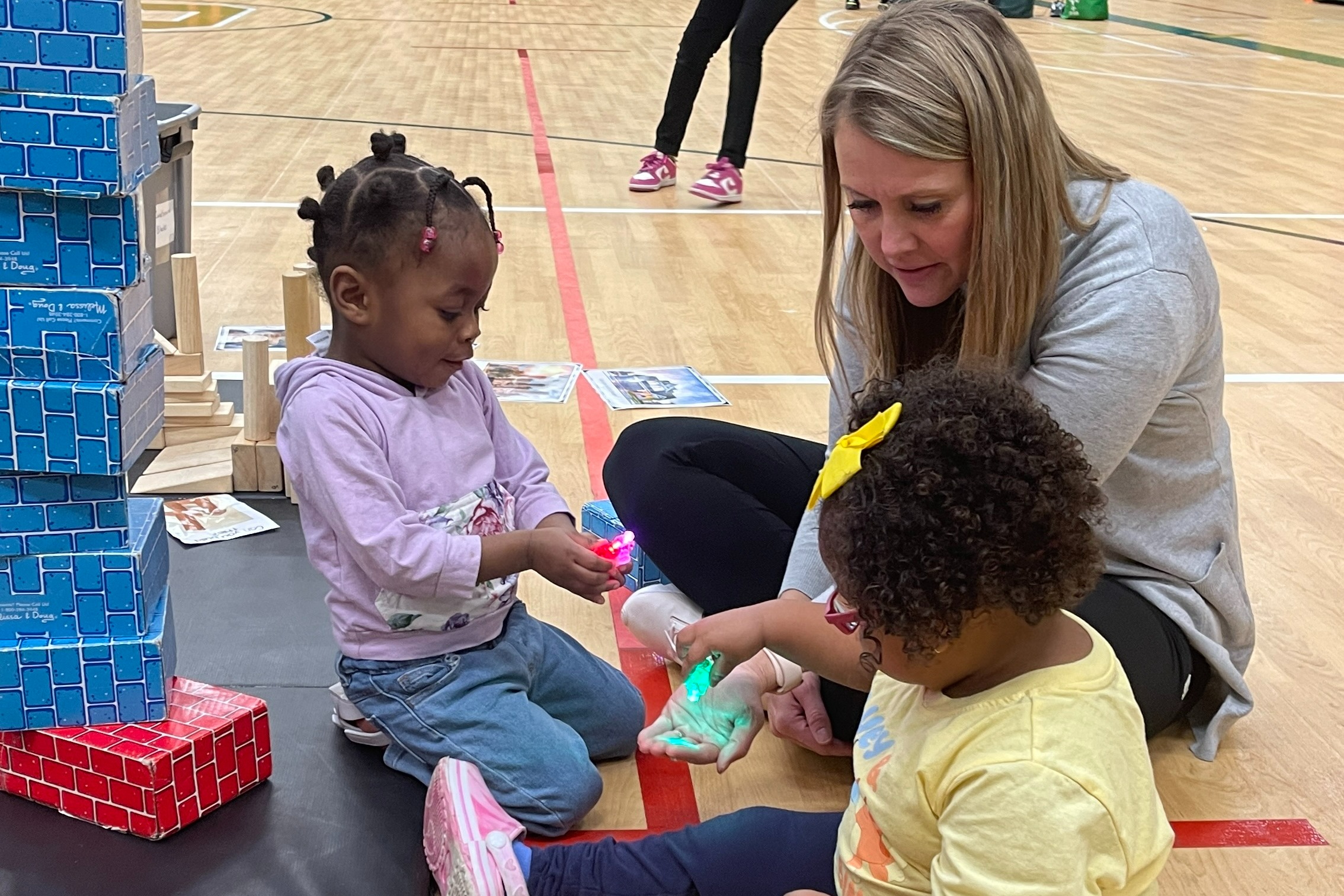 Two children and a teacher play with blocks on the floor.