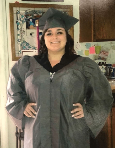 A woman wearing a graduation gown stands in a kitchen, symbolizing success.