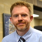 A bearded man in a tie standing in front of a wall.