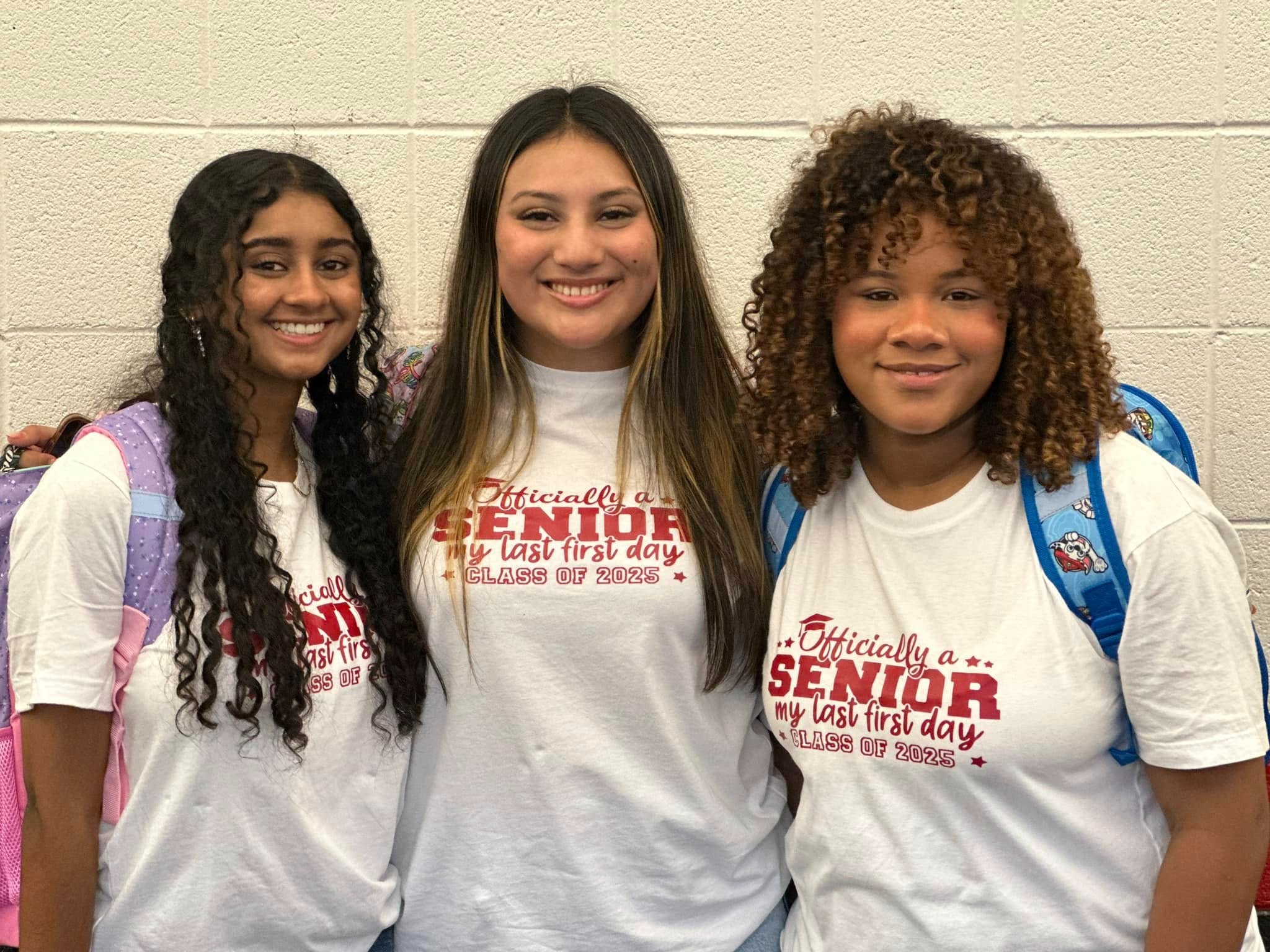 Three high school senior girls smiling and posing together in a school hallway. They are wearing matching white T-shirts with red text that reads, 'Officially a Senior – My Last First Day – Class of 2025.' Each student has a backpack, and they stand closely with their arms around each other, expressing excitement for their senior year.