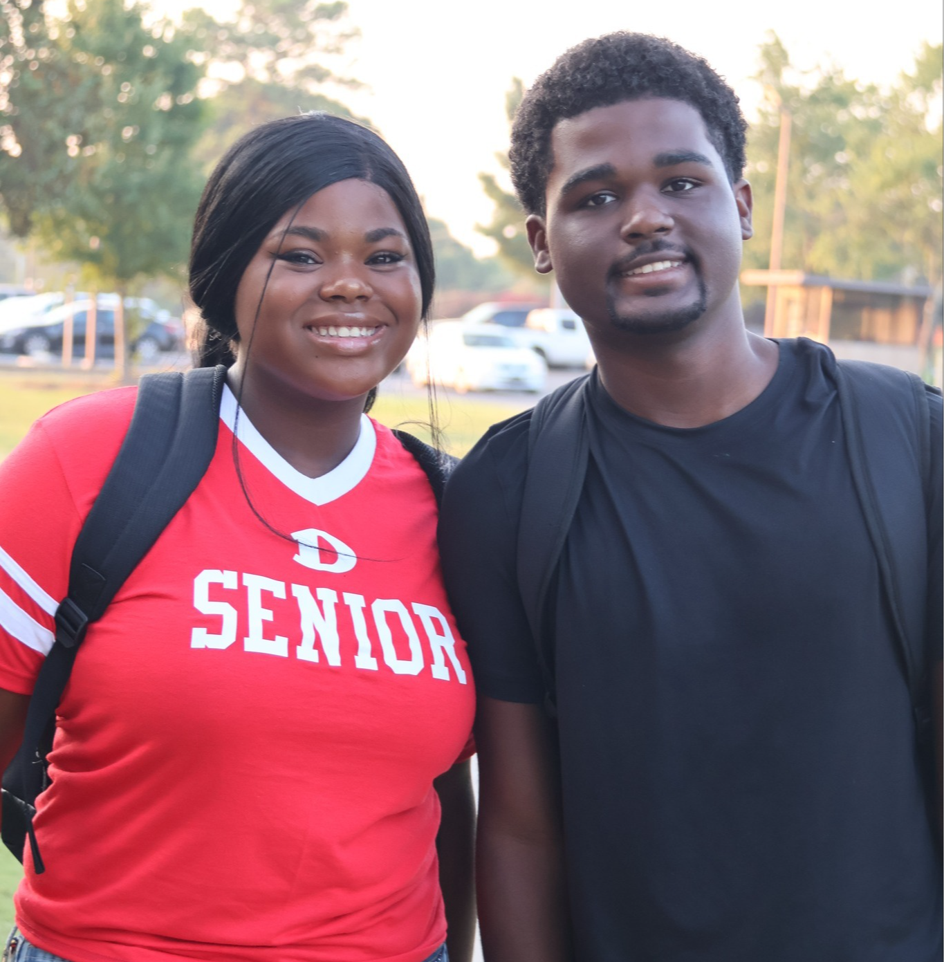 Two high school seniors smiling outdoors on campus. The student on the left is wearing a red and white 'Senior' T-shirt with a large 'D' logo, along with decorated jeans featuring glittery letters. The student on the right is dressed in a black T-shirt. Both are carrying backpacks and standing close together, looking happy as they start their senior year. Cars and trees are visible in the background, indicating a school setting.