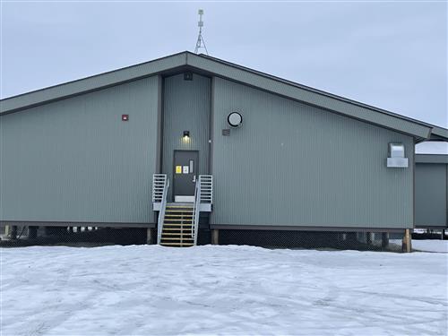 The Image shows a large building with a door and stairs leading to it. The building is surrounded by snow and there is a lot of snow on the ground.  According to a Google Search, the building is the Allakaket School . It is a school located in Allakaket, Alaska.