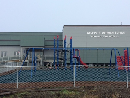 The building itself is a large red brick building with a black metal fire escape on the side. In front of the building is a playground with a green metal fence surrounding it. A sign above the main entrance reads "Andrew K. Demoski School - Home of the Wolves".